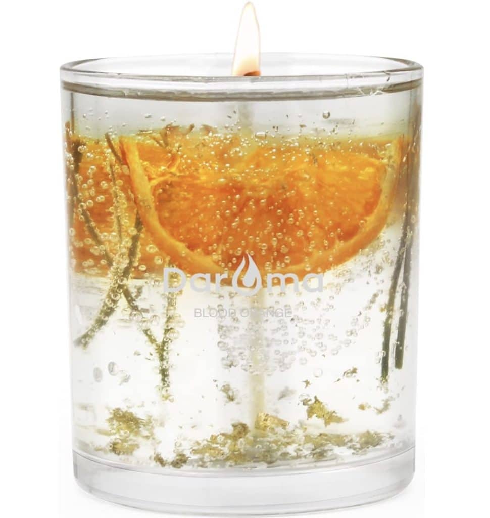 A clear candle encased in a glass container showcases suspended slices of blood orange and sprigs of greenery, resembling the delicate charm of candles with dried flowers. The candlewick is lit, casting a warm glow from the small flame visible at the top.